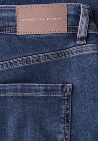 Jean en denim bleu foncé avec une étiquette en cuir portant la mention « STREET ONE STUDIO ». Dispose de coutures décoratives et d'un design visible des poches arrière.