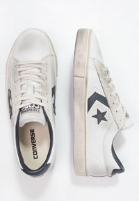 Baskets Converse en blanc cassé et noir. Comprennent une tige en cuir lisse, un bout arrondi, une fermeture à lacets, et un logo étoile contrastant.