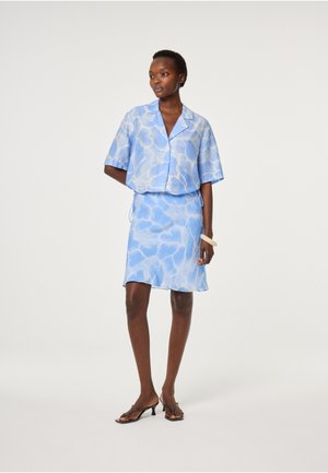 Vrouw draagt een blauw, kort mouwig, gebloemd blousehemd met knopen en een bijpassende knielange rok, staand met één hand achter haar rug.