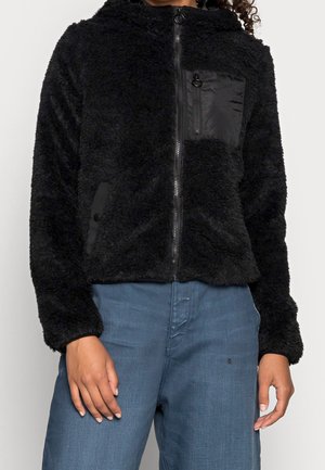 Personne portant une veste noire duveteuse zippée avec une poche poitrine zippée et un jean bleu, debout, mains détendues.