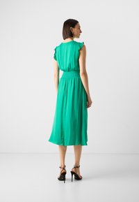 Green dress with short ruffled sleeves, smocked waist, flowy skirt, and ankle-length design. Worn with black high-heeled sandals.