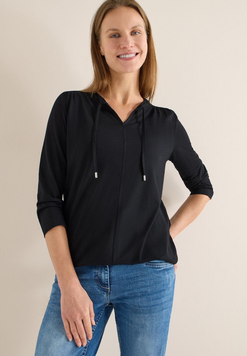 Haut noir à capuche avec col en V, manches trois-quarts et cordons de serrage, associé à un jean skinny bleu. Tissu lisse et coupe décontractée.