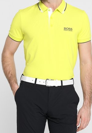 Mand iført en knaldgul Hugo Boss polo, stoppet ned i sorte bukser med et hvidt bælte, hænder hvilende nær lommerne.