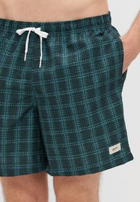 Calções de banho pretos com um padrão quadriculado em azul-piscina, cintura elástica, cordão branco e bolsos laterais. Tecido texturizado, vestuário de verão.