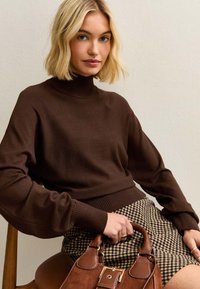 Maglione a maglia marrone con colletto alto e maniche lunghe, abbinato a una gonna a quadri. Una borsa marrone è tenuta in mano, su una sedia di legno.