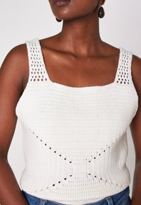 Femme portant un haut sans manches en tricot blanc avec un décolleté carré et des détails à motifs, associé à un jean bleu et des boucles d'oreilles ovales foncées.