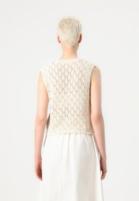 Knit sleeveless top in beige with a diamond pattern. Textured fabric and open design. Straight hem at the back, paired with a light skirt.