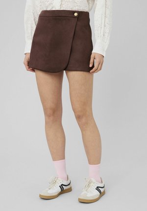 Mini-jupe portefeuille marron avec bouton doré, portée avec un chemisier blanc à œillets, des chaussettes roses et des baskets blanches à accents noirs.