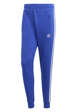 Mann trägt einen blauen Adidas Trainingsanzug mit weißen Streifen an Ärmeln und Hosen, steht mit einer Hand in der Tasche und weißen Sneakers.