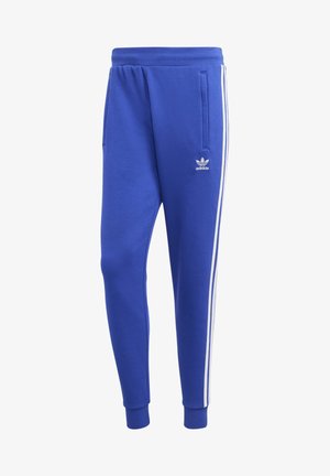 Mann trägt einen blauen Adidas Trainingsanzug mit weißen Streifen an Ärmeln und Hosen, steht mit einer Hand in der Tasche und weißen Sneakers.