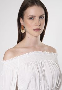 Weißen Off-the-Shoulder-Oberteil mit gesmocktem Ausschnitt, versehen mit zarten gestickten Details. Holz- und geflochtene Ohrringe sorgen für texturierte Akzente.