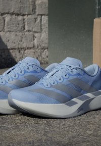 Jasnoniebieskie buty do biegania z szarymi paskami i grubymi białymi podeszwami, spoczywające na utwardzonej powierzchni na tle kamiennego muru.
