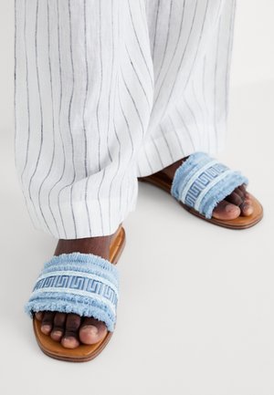 Niebieskie klapki z materiału z postrzępionymi krawędziami i wzorzystymi paskami, uzupełnione brązową, płaską podeszwą. Widoczne są białe spodnie w paski.