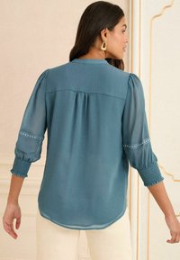 Blusa verde acqua con retro plissettato, maniche puff trasparenti con polsini elastici e decorazioni vicino ai polsini. Tessuto leggero.