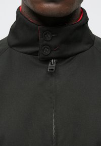 Sort jakke med høj krave og rød indvendig foring. Har to sorte knapper og lynlås til lukning. Glat tekstur og skræddersyet design.