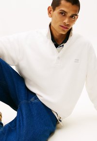 Λευκή φλις μπλούζα με όρθιο γιακά, με δύο κουμπιά και μικρό γκρι λογότυπο. Το σύνολο ολοκληρώνεται με ένα ζευγάρι μπλε τζιν παντελόνι.