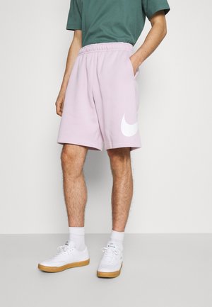 Muž nosící světle fialové šortky Nike s bílým logem, zelené tričko, bílé ponožky a bílé tenisky s gumovou podrážkou.