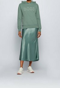 Vihreä puuvillapaita "BOSS" -logolla, yhdistettynä kiiltävään vihreään satiiniseen midi-hameeseen ja vaaleisiin tennareihin, joissa on vaaleanpunaisia yksityiskohtia.