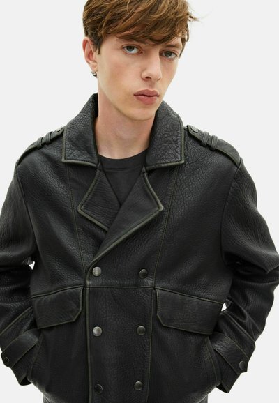 Chaqueta de cuero negra con doble botonadura, grandes solapas, acabado texturizado, acentos de costura verde y detalles de botones en los bolsillos y hombros.