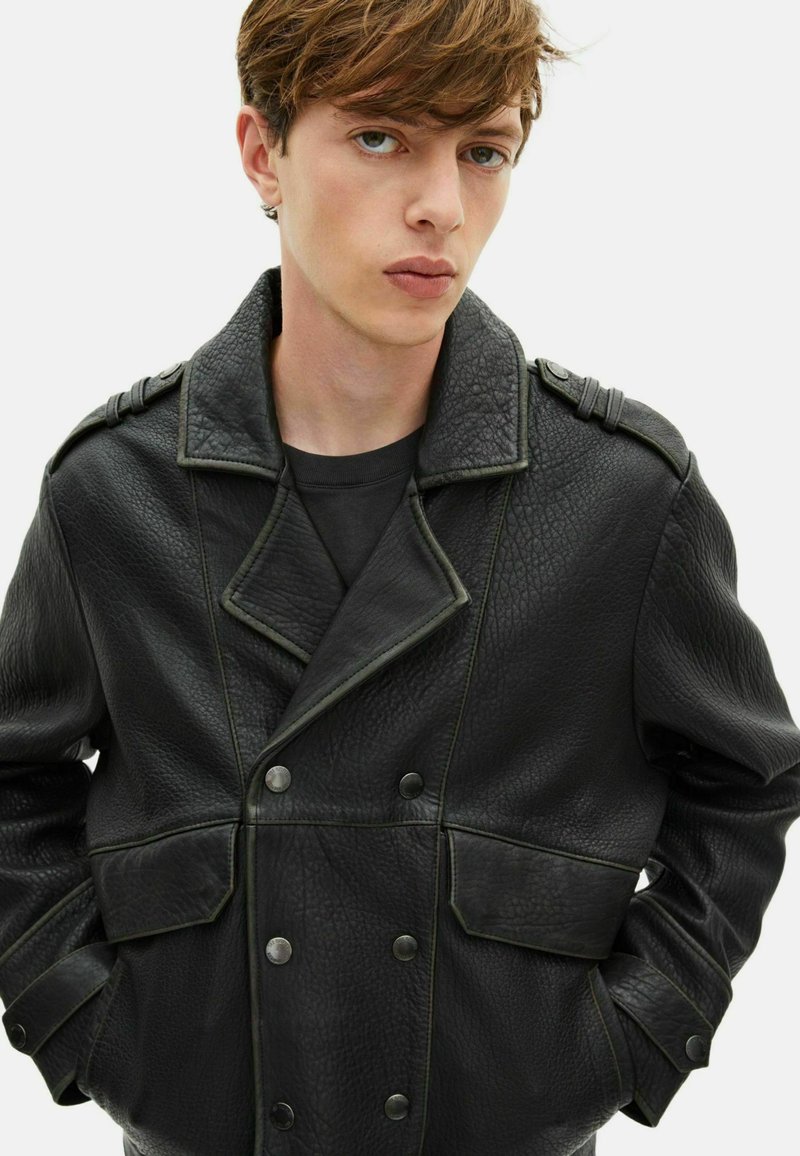 Veste noire en cuir à double boutonnage avec grands revers, finition texturée, accents de couture verts et détails de boutons sur les poches et les épaules.