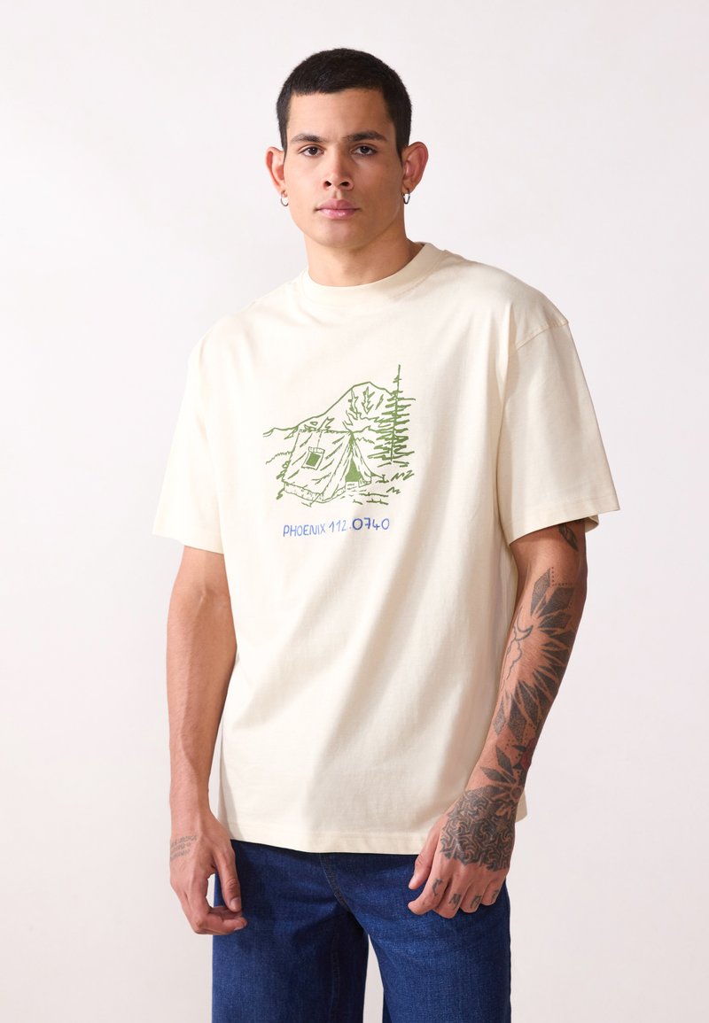 Camiseta de algodón cremosa con un gráfico verde de exterior, "PHOENIX 112.0740", y un corte relajado. Llevada con unos jeans de mezclilla azules.