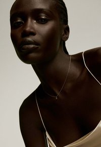 Collar con cruz de oro con una cadena fina, lucido sobre una textura de piel oscura y suave. La figura está vestida con una blusa de satén ligera con tirantes delgados.