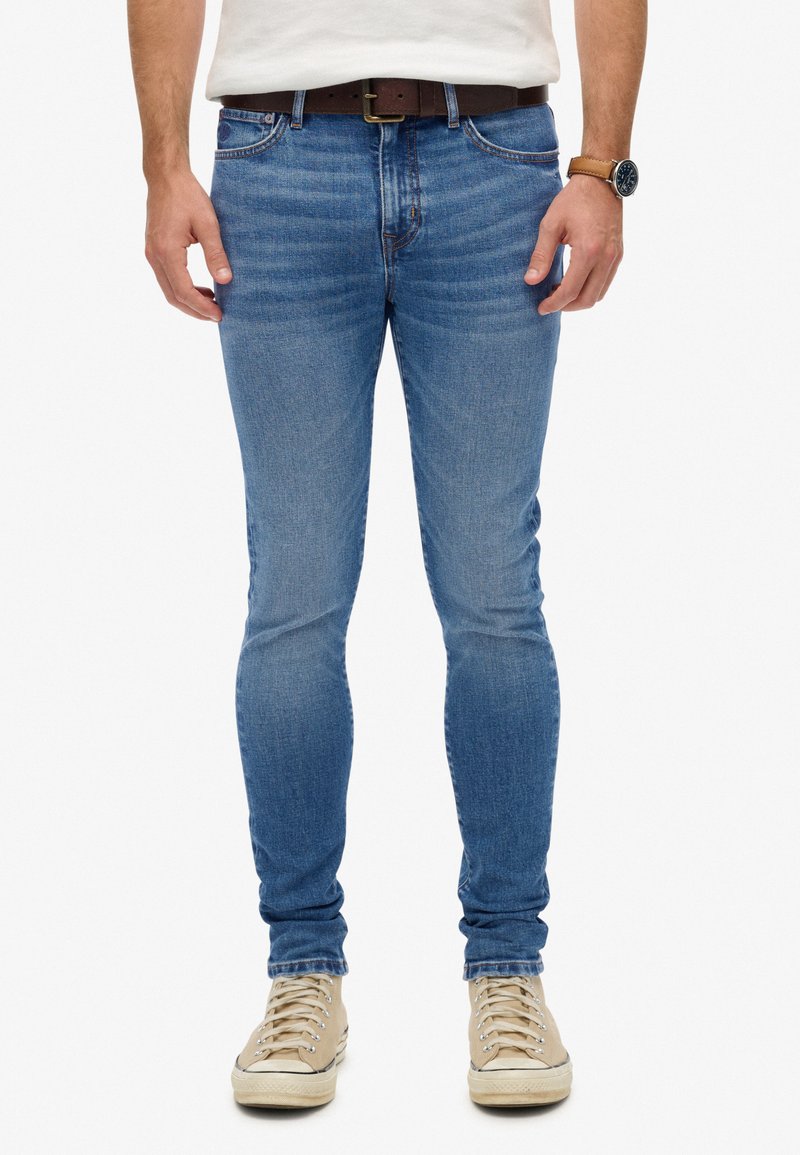 Jeans skinny azul desgastados con una textura suave, diseño de cinco bolsillos, costuras sutiles, llevados con un cinturón marrón y zapatillas beige.