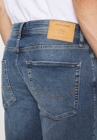 Vaqueros de mezclilla azul con un ligero desvanecimiento. Presentan costuras amarillas y una etiqueta marrón en la cinturilla. Dos bolsillos traseros con costuras curvas.