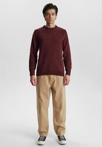 Burgunderfarbener Strickpullover mit gerippten Bündchen und Saum, kombiniert mit beigen, geraden Hosen und schwarzen Low-Top Sneakers.