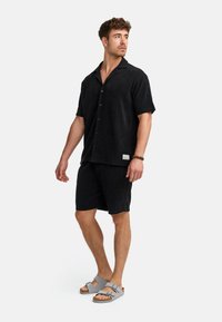 Sort kortærmet skjorte med krave, parret med sorte shorts. Sættet har struktureret stof og lysegrå sandaler.