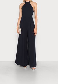 Marinblå jumpsuit med hög halterneck, åtsittande liv och vida ben. Slätt tyg, veck vid midjan, kombineras med en metallisk clutch.