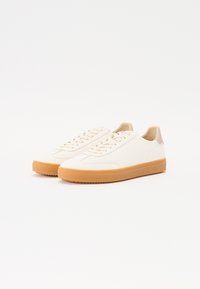 Sneakers bianchi in pelle con un tallone beige e una suola in gomma color nocciola. Presentano un design minimalista e una chiusura con lacci classica.