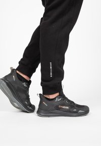 Zwarte sportieve schoenen met een gestructureerde mesh bovenkant, grijze accenten en een gewatteerde kraag, gecombineerd met zwarte tapered joggingbroeken met witte tekst.