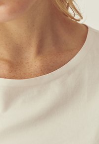 T-shirt en coton blanc avec un large col arrondi. Le tissu semble lisse avec un léger éclat. La peau parsemée de taches de rousseur est visible.
