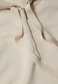 Sweat à capuche zippé beige en tissu doux, avec une fermeture éclair proéminente et des cordons épais. Les coutures donnent de la structure au design.