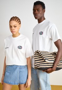 Duas pessoas usam camisetas brancas com um logo colorido de "WORLD TOUR". A mulher veste uma saia de jeans; o homem segura uma bolsa de palha listrada.