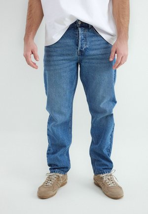 Osoba oblečená v modrých džínsach, béžových teniskách s bielymi šnúrkami a bielou košeľou stojí na jednoduchom svetlom pozadí.