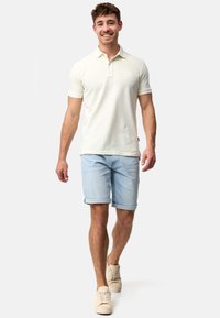 Creme polo-shirt med korte ærmer, der har et diskret logo. Sammen med lyseblå opsmøgede shorts og beige sneakers.