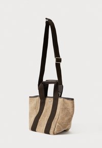 Beżowa torba tote z teksturowanej tkaniny z ciemnobrązowymi pionowymi paskami i regulowanym paskiem na ramię na jednolitym tle.
