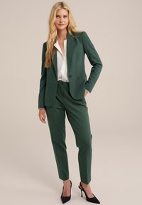 Completo verde su misura con giacca a un bottone e pantaloni slim-fit, abbinato a una camicia bianca e scarpe con tacco nero.