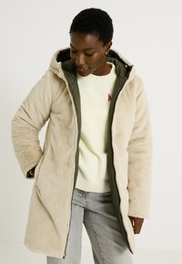 Chaqueta de forro polar beige con un forro verde oliva con cremallera, que incluye capucha y bolsillos laterales. Textura suave con un ajuste relajado.