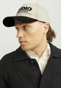 Beige kasket med sort skygge, med broderet tekst. Modellen bærer en struktureret sort jakke over en hvid skjorte med knapkrave.