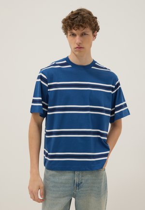 Giovane uomo con capelli ricci che indossa una maglietta a righe orizzontali blu e bianca e jeans azzurri, in piedi davanti a uno sfondo semplice.