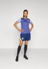 Atlético Madrid fotbaluniform med en lilla topp med gule detaljer, marineblå shorts og sorte sokker, supplert med sorte fotballsko.