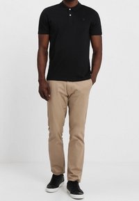 Homme portant un polo noir à manches courtes, un pantalon beige et des baskets noires avec semelles blanches, se tenant devant un fond uni.