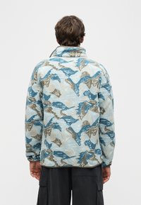 Άνδρας που φοράει φλις μπουφάν με αφηρημένο σχέδιο σε ανοιχτό μπλε, μπεζ και καφέ αποχρώσεις, από πίσω, συνδυασμένο με σκουρόχρωμο παντελόνι cargo.