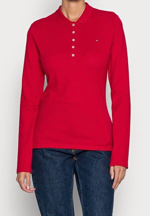 Kvinde iført en rød langærmet polo med fem knapper kombineret med mørkeblå jeans, stående mod en ensfarvet baggrund.