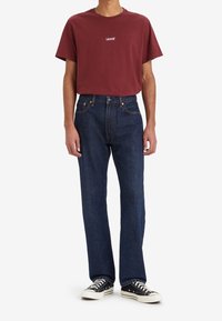 Kastanjefärgad t-shirt med kort ärm och "LEVI'S"-logga, i kombination med mörkblå raka jeans och svarta låga sneakers, utrustade med vita snören.