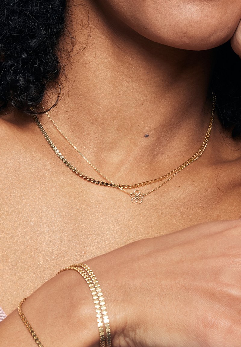 Złote naszyjniki warstwowe z płaskim łańcuchem i delikatnym wisiorkiem w kształcie kwiatu; bransoletki z połączonym złotym wzorem, wyróżniające się wypolerowanym wykończeniem.