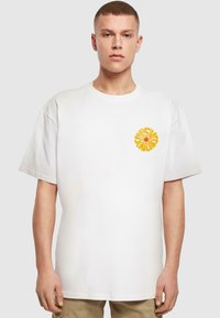 Hvid bomulds-t-shirt med korte ærmer og en afslappet pasform. Har et klart gult rundt grafisk motiv på venstre brystområde.